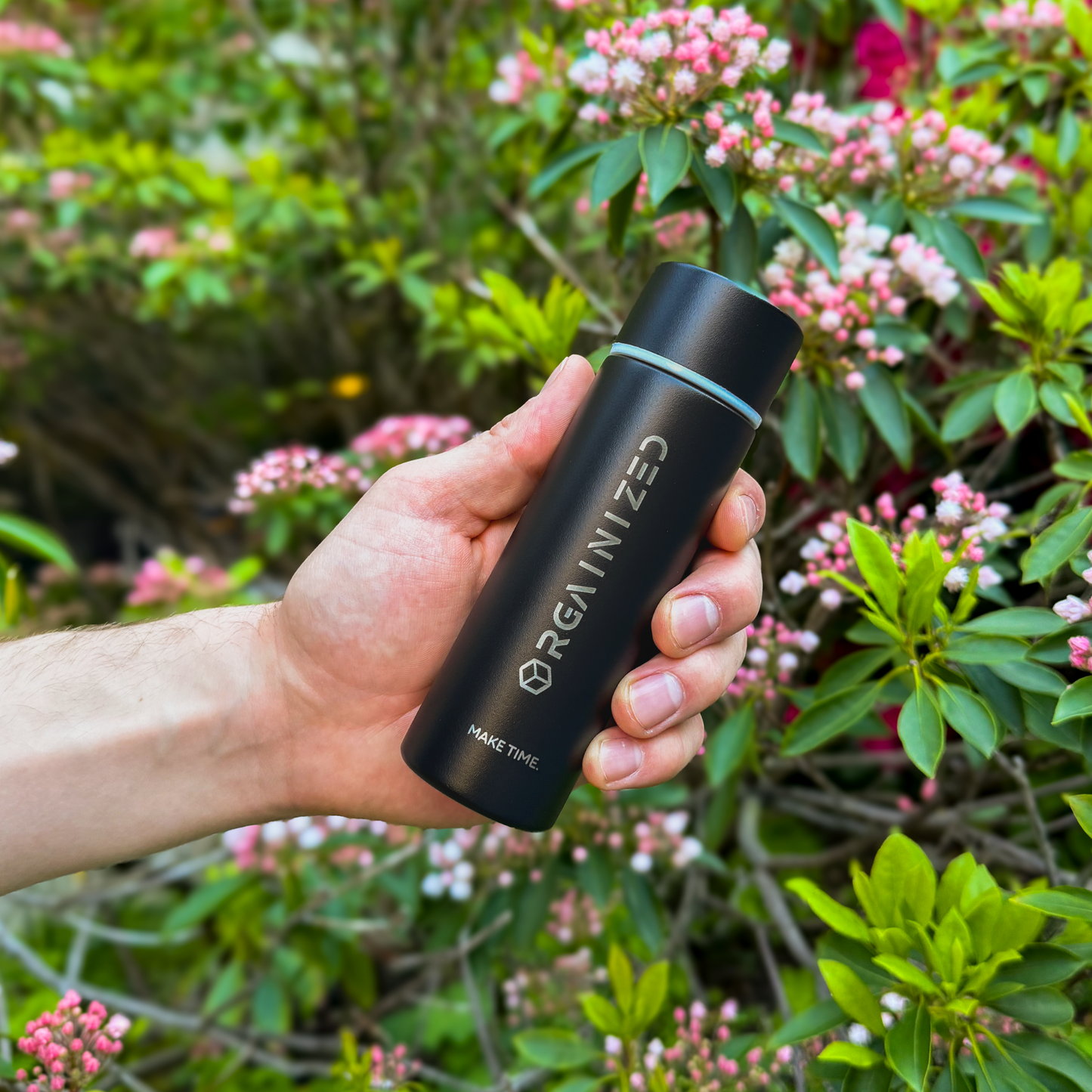 matte_black_2 – angled view. Black cylindrical container with 'Orgainized' branding being held at a tilt in front of a flowering bush