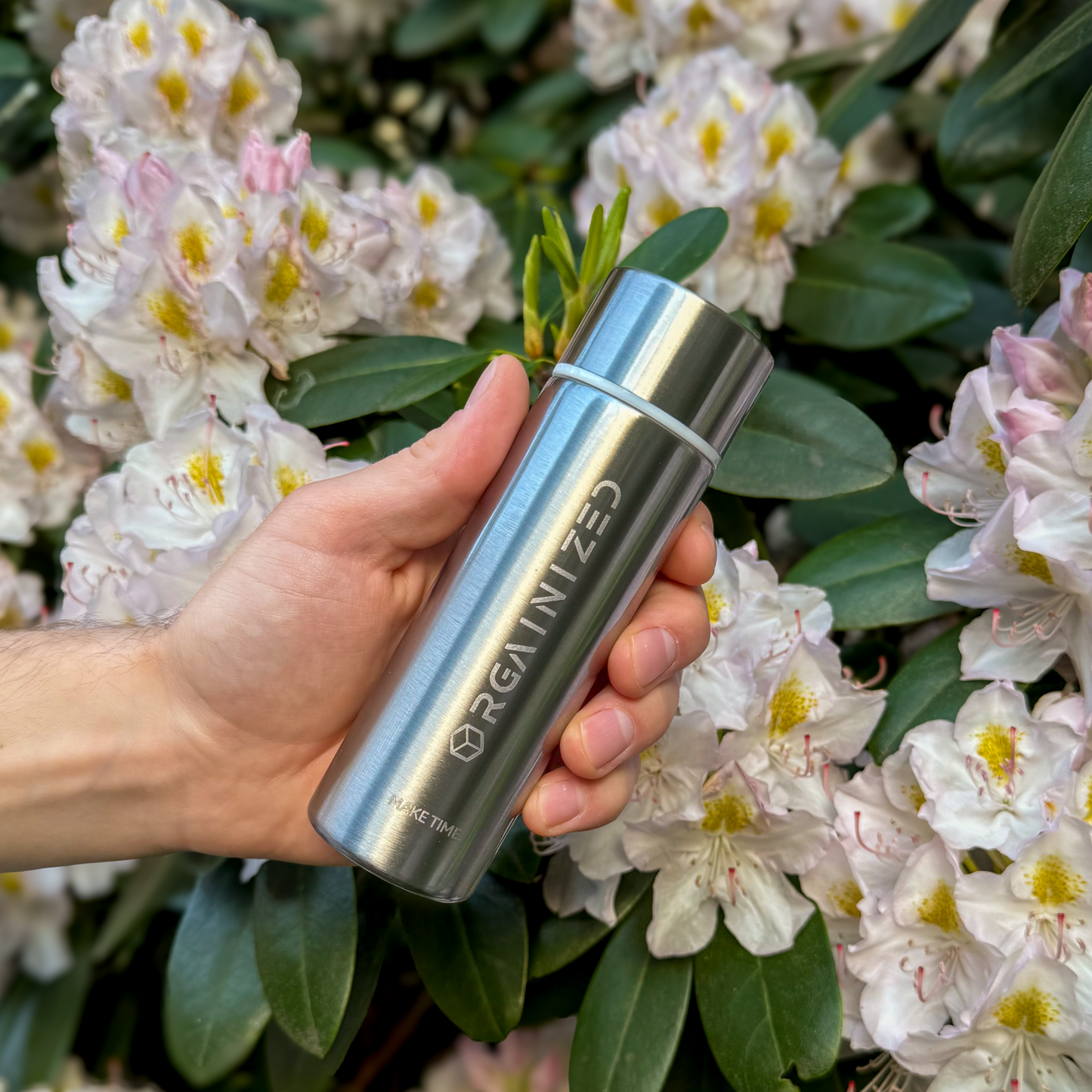 natural_steel_2 – angled view. Silver cylindrical container with 'Orgainized' branding being held at a tilt in front of a flowering bush