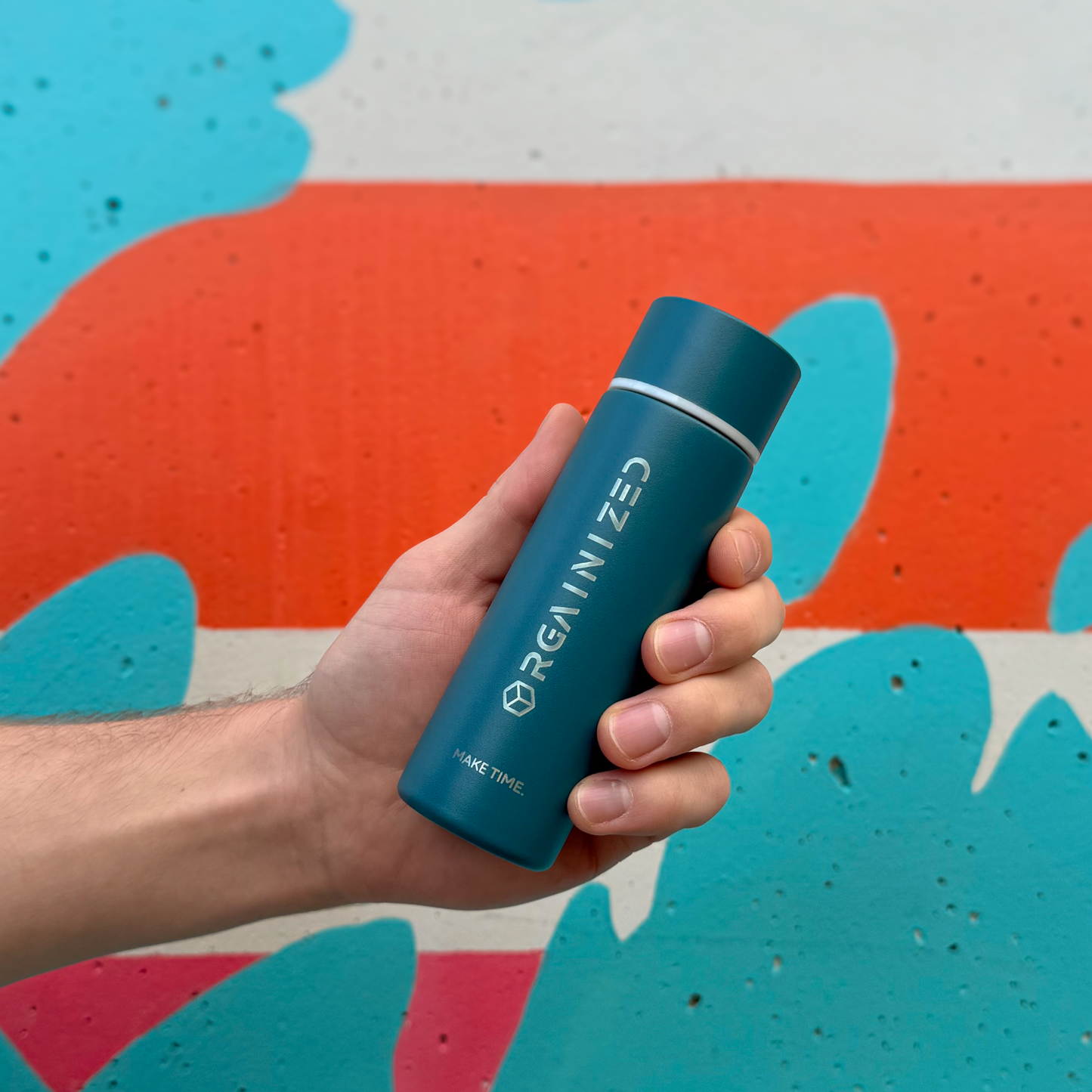 rogue_blue_2 – angled view. Blue/teal cylindrical container with 'Orgainized' branding being held at a tilt in front of a painted wall