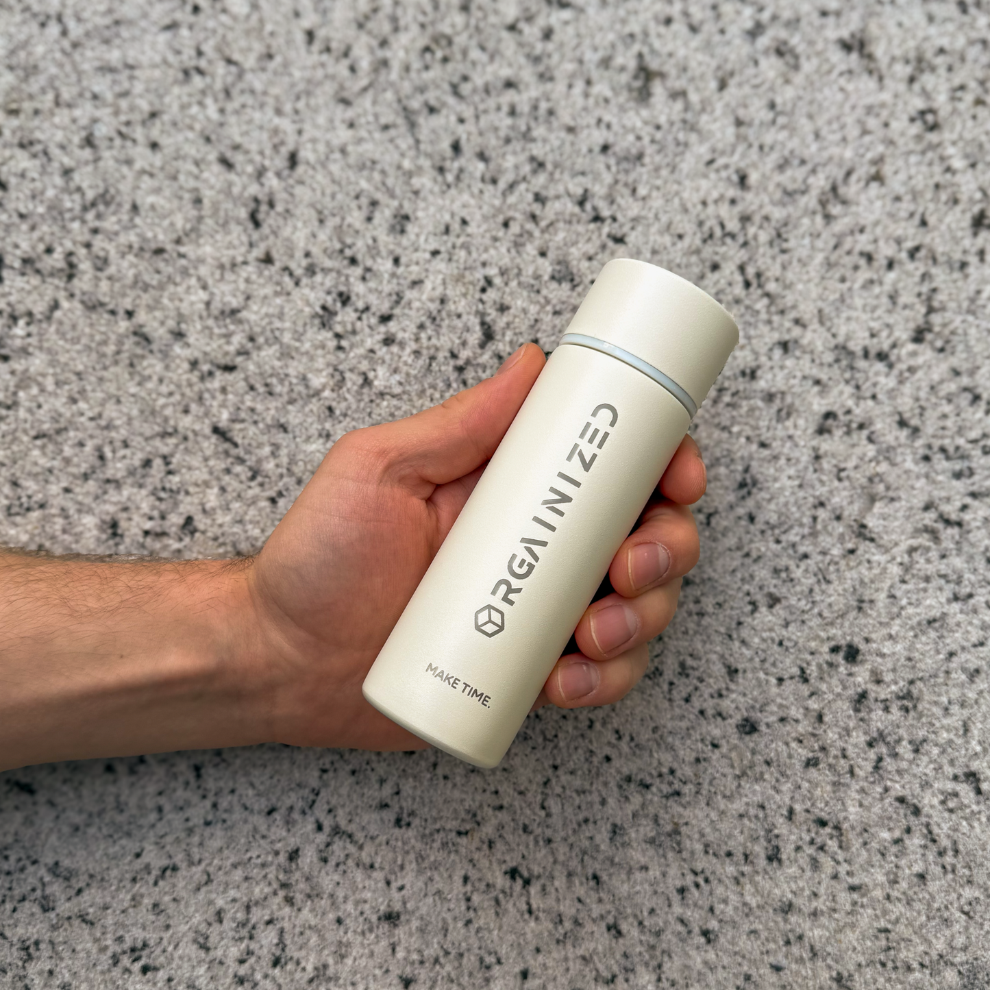 white_sand_2 – angled view. Off-white cylindrical container with 'Orgainized' branding being held at a tilt in front of a granite wall