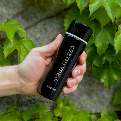 gloss_black_2 – angled view. Black cylindrical container with 'Orgainized' branding being held at a tilt in front of a vine-covered wall