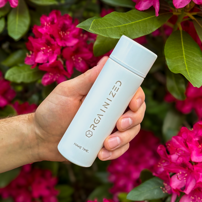 gloss_white_2 – angled view. White cylindrical container with 'Orgainized' branding being held at a tilt in front of a flowering bush